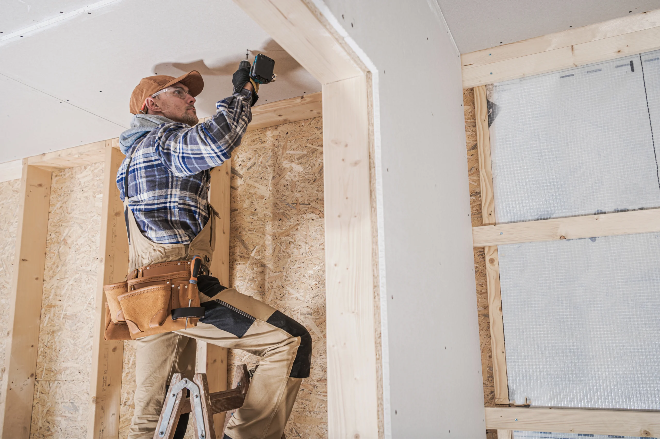 Man wearing ballcap and safety googles on ladder with tool belt drilling a screw in the ceiling